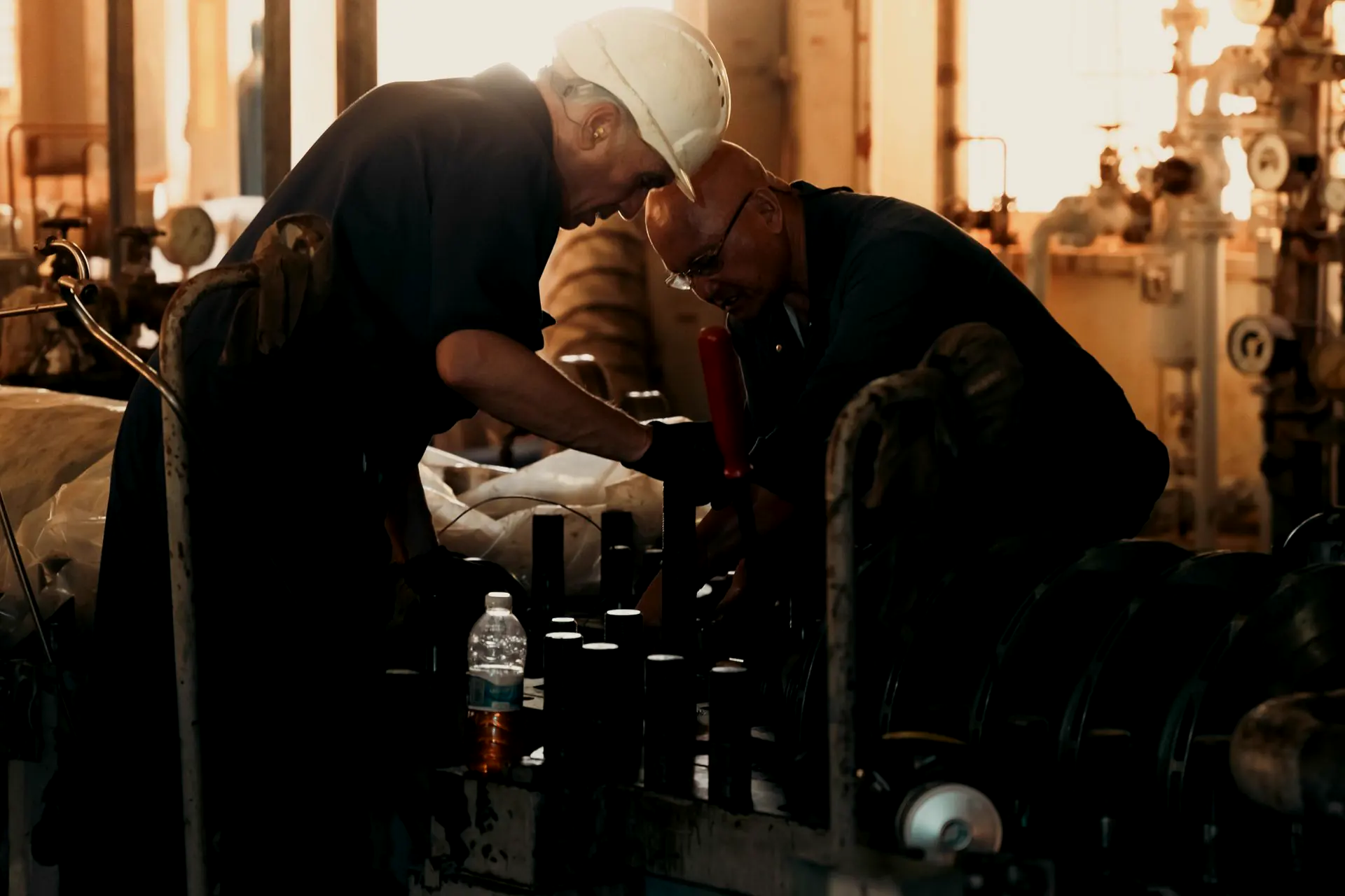 Two tradesmen in dark coveralls, one in a white hard hat, working together over a row of machinery studs in a plant, warm light behind them